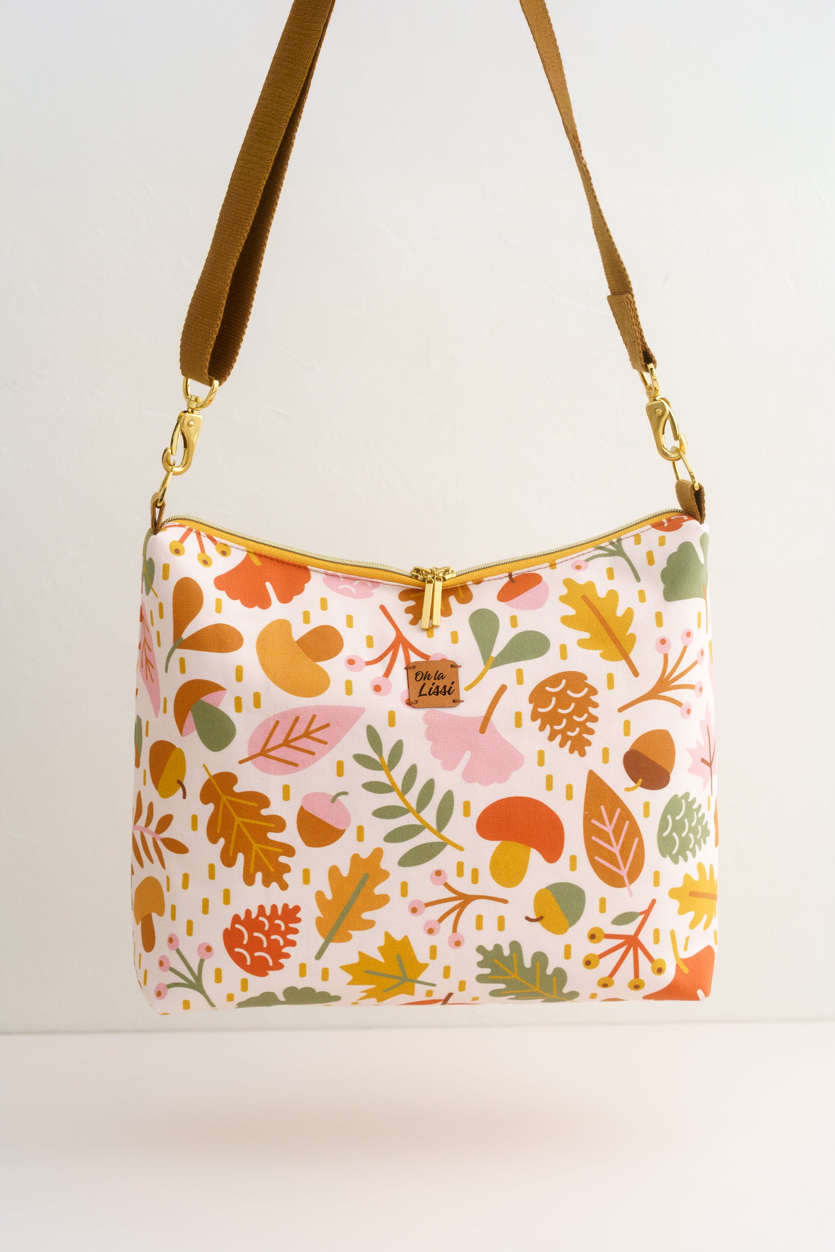 Eine Umhängetasche mit einem farbenfrohen Herbstmuster aus Blättern, Eicheln und Pilzen. Die Tasche hat einen braunen Riemen, goldfarbene Beschläge und einen goldenen Reißverschluss. Der Text "Oh la Lissi" befindet sich auf der Tasche.